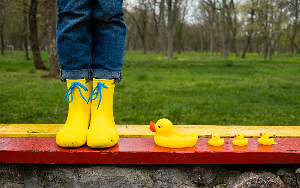 A child in bright yellow boots stands next to a mother and chicks set of rubber duckies