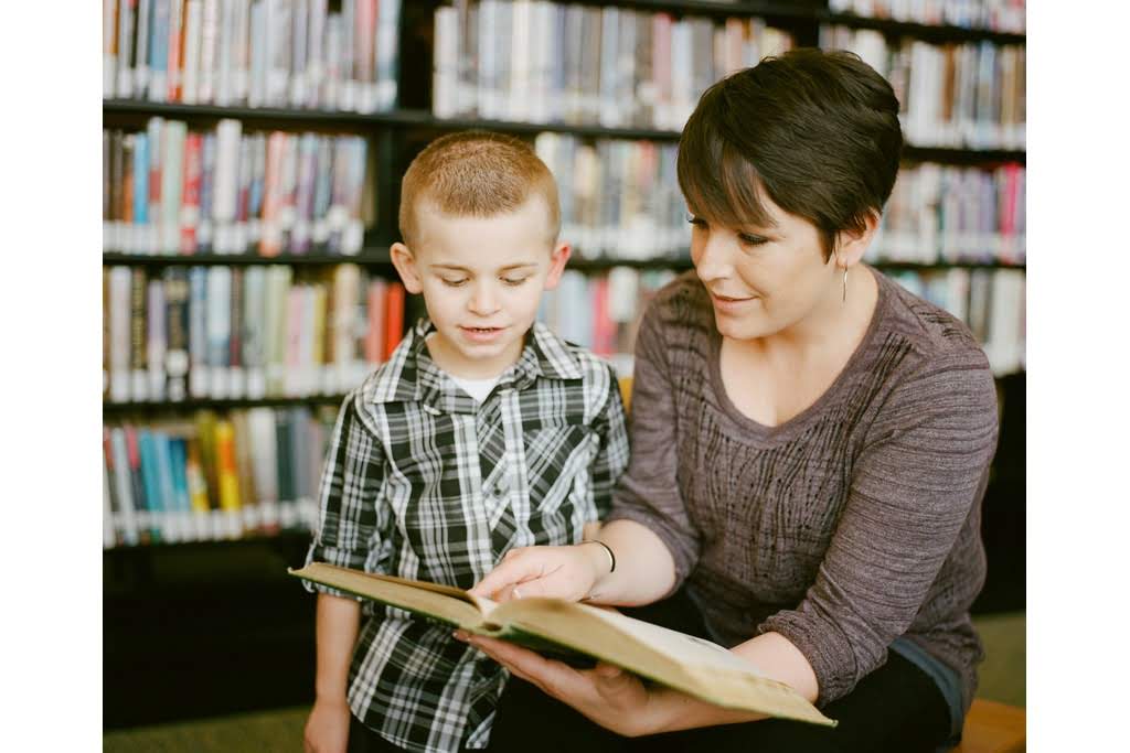 A Caucasian woman and boy looking at a book together in a library teacher teaching student to read