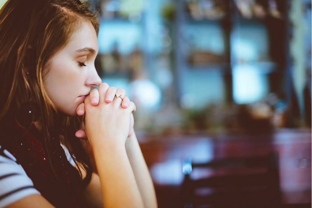 A Caucasian woman clasping her hands together with her eyes closed woman prayinhg