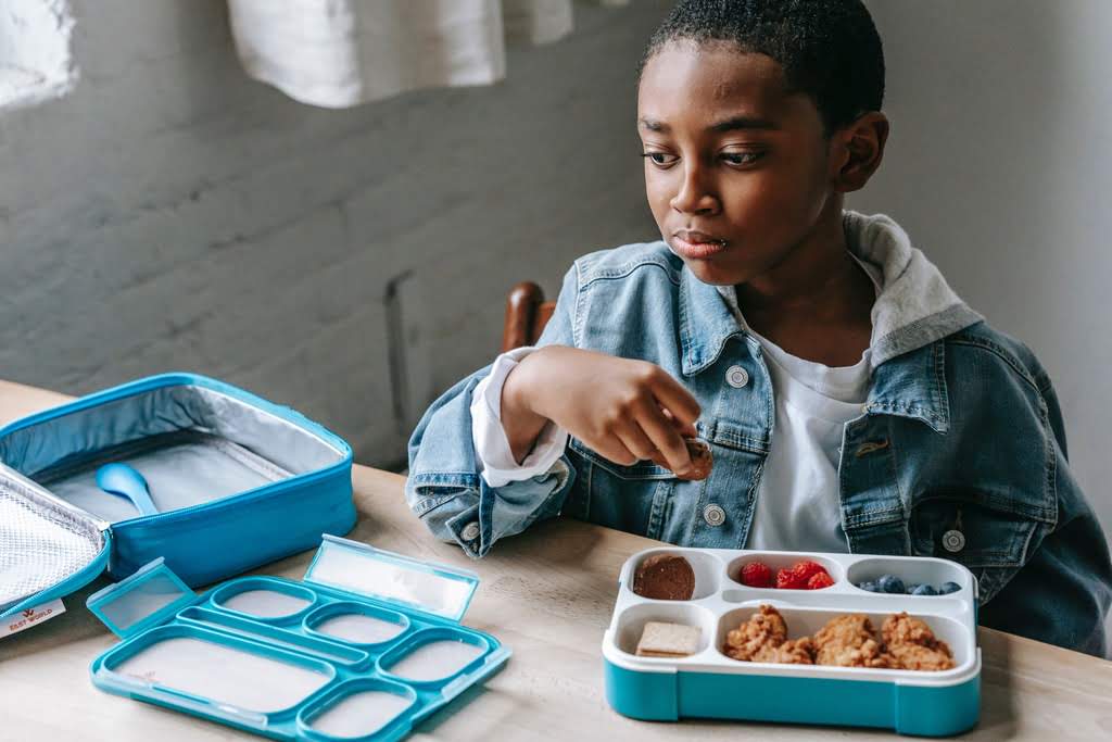 A Black boy eating lunch from a lunchbox at a table kid eating lunch