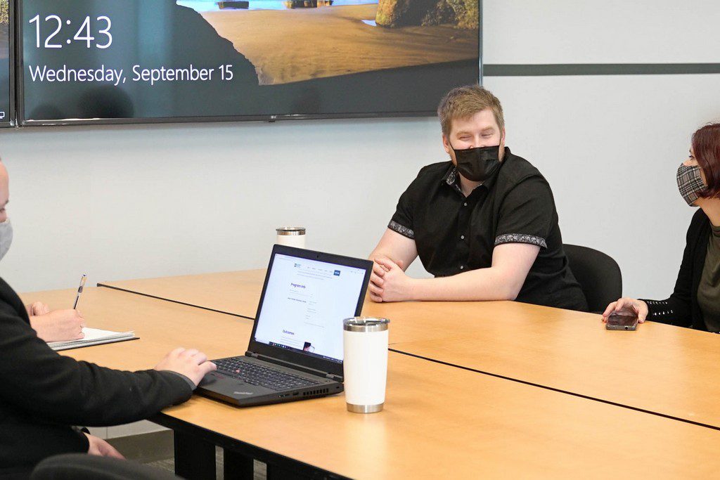 Three White individuals with masks sit around a table and discuss team meeting in the boardroom