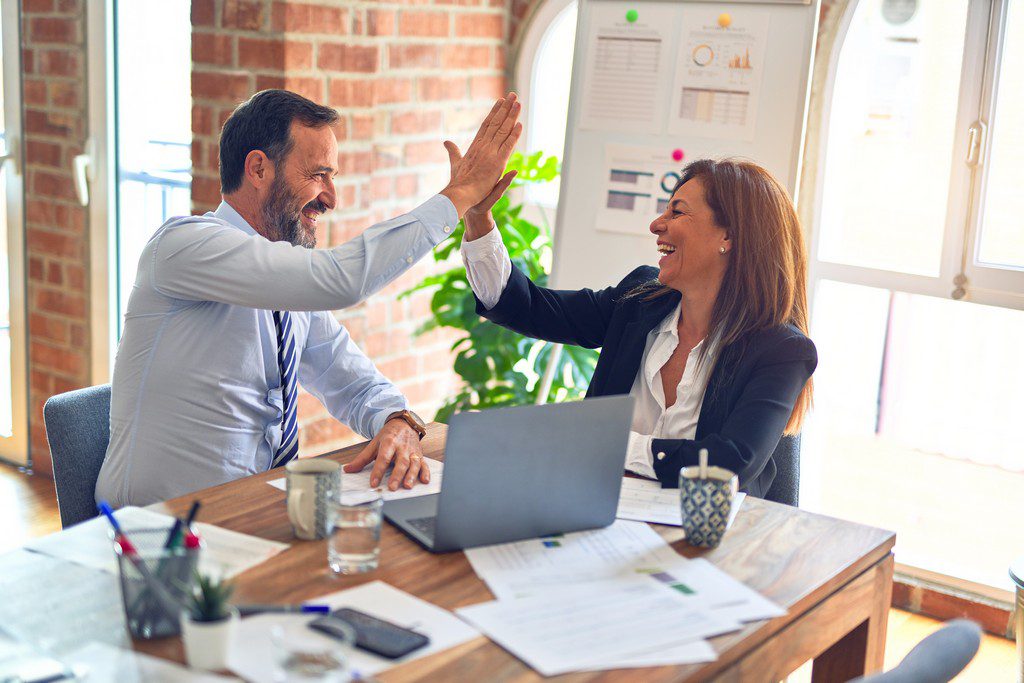 An older white man and white woman in business attire high five each other while laughing work team celebrating