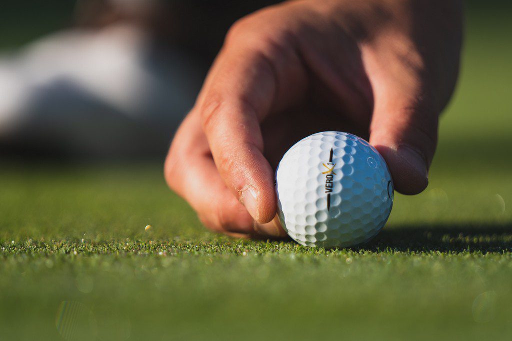 A person places a gold bowl on a lawn man teeing up his golf ball